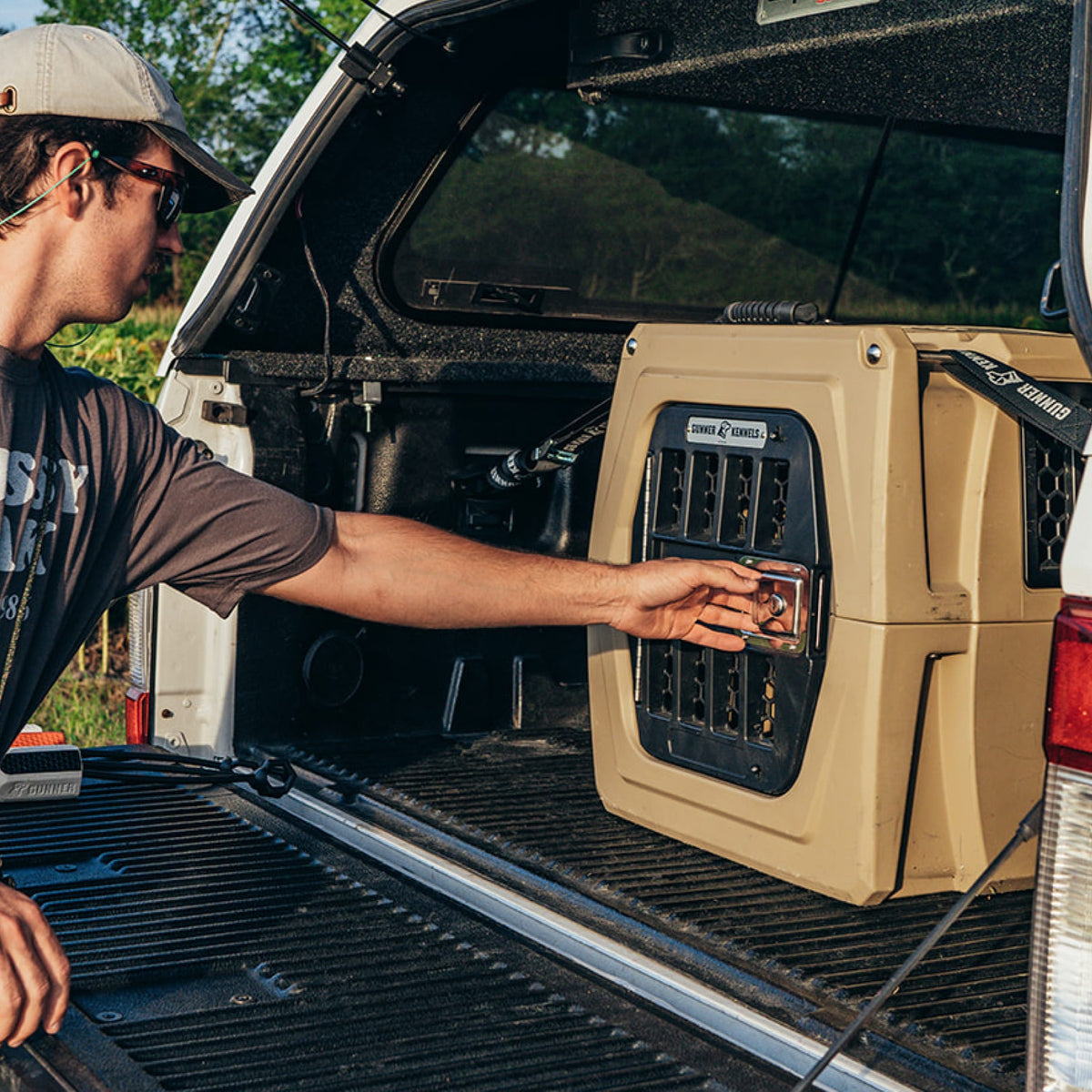 A person in sunglasses opens a GUNNER G1™ KENNEL - TAN, the safest double-walled dog crate, secured in the back of a pickup truck with a black bed liner, outdoors on a sunny day.