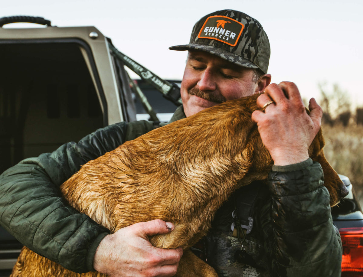 A man wearing a camouflage hat and jacket hugs his brown working dog tightly outdoors near a vehicle, showing affection and a close bond.