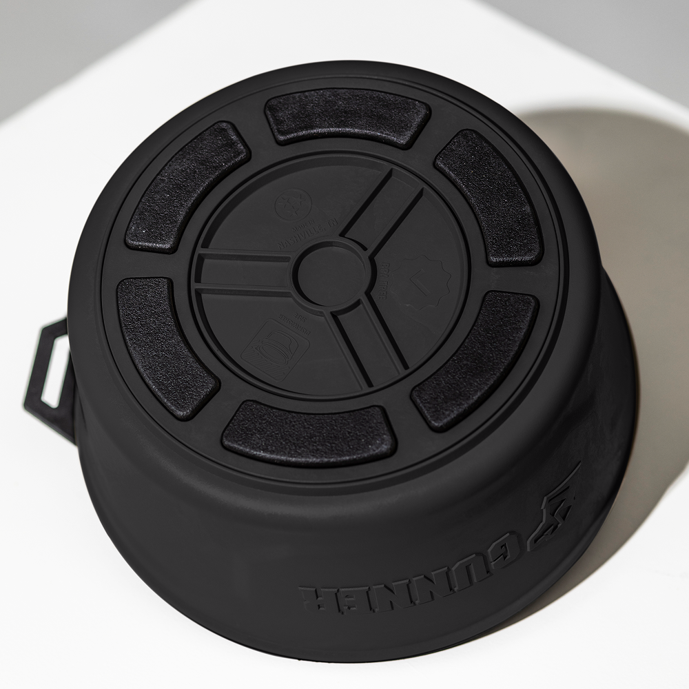 A black, round GUNNER DOG BOWL by GUNNER with a textured, segmented lid is placed upside down on a white surface. GUNNER and an animal logo are embossed on the side.