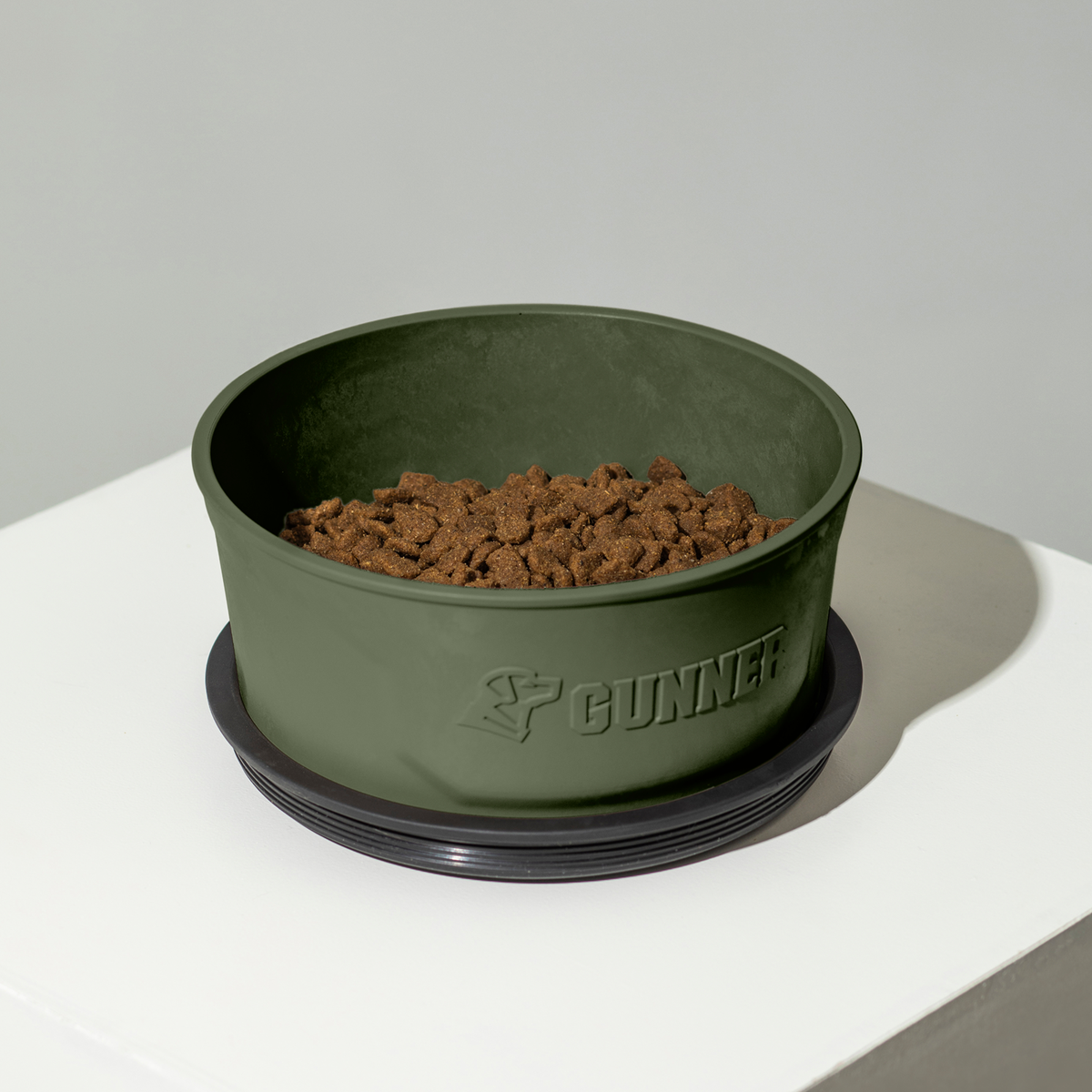A GUNNER DOG BOWL - GUNNER GREEN by GUNNER, made in America and filled with dry kibble, sits on a black non-slip base on a white surface against a light gray background.