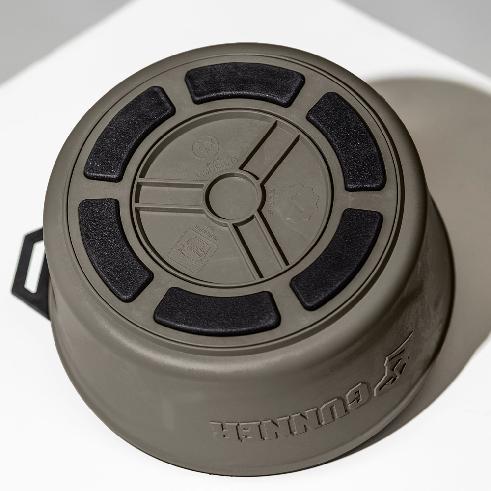 A gunmetal GUNNER DOG BOWL with black rubber grips on the base sits upside down on a white surface. The brand name GUNNER is embossed on the side of this portable dog bowl.