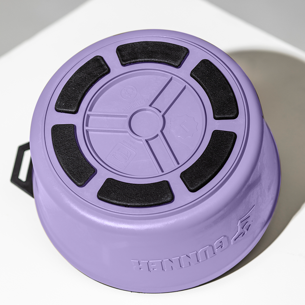The limited-edition GUNNER DOG BOWL - LAVENDER by GUNNER is shown upside down, revealing its circular base with black non-slip pads, embossed symbols, a matte finish, and a black side handle for added style and function.