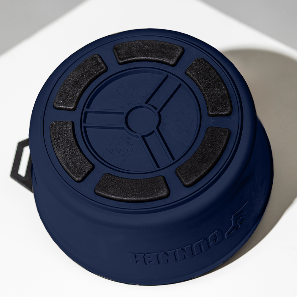 A limited-edition GUNNER DOG BOWL - NAVY by GUNNER is shown upside down on a white surface, displaying black non-slip grips, embossed symbols on the bottom, and a handle on one side.