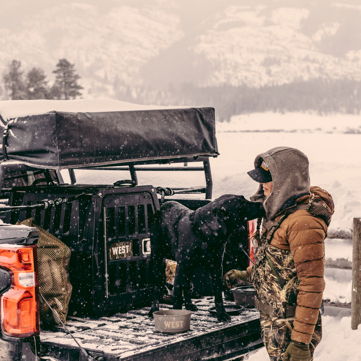 Dressed in winter gear, a person stands by a truck in snowy mountains, interacting with a black dog inside the GUNNER G1™ KENNEL - BLACK crate in the truck bed. Snow falls as supplies sit visible nearby.