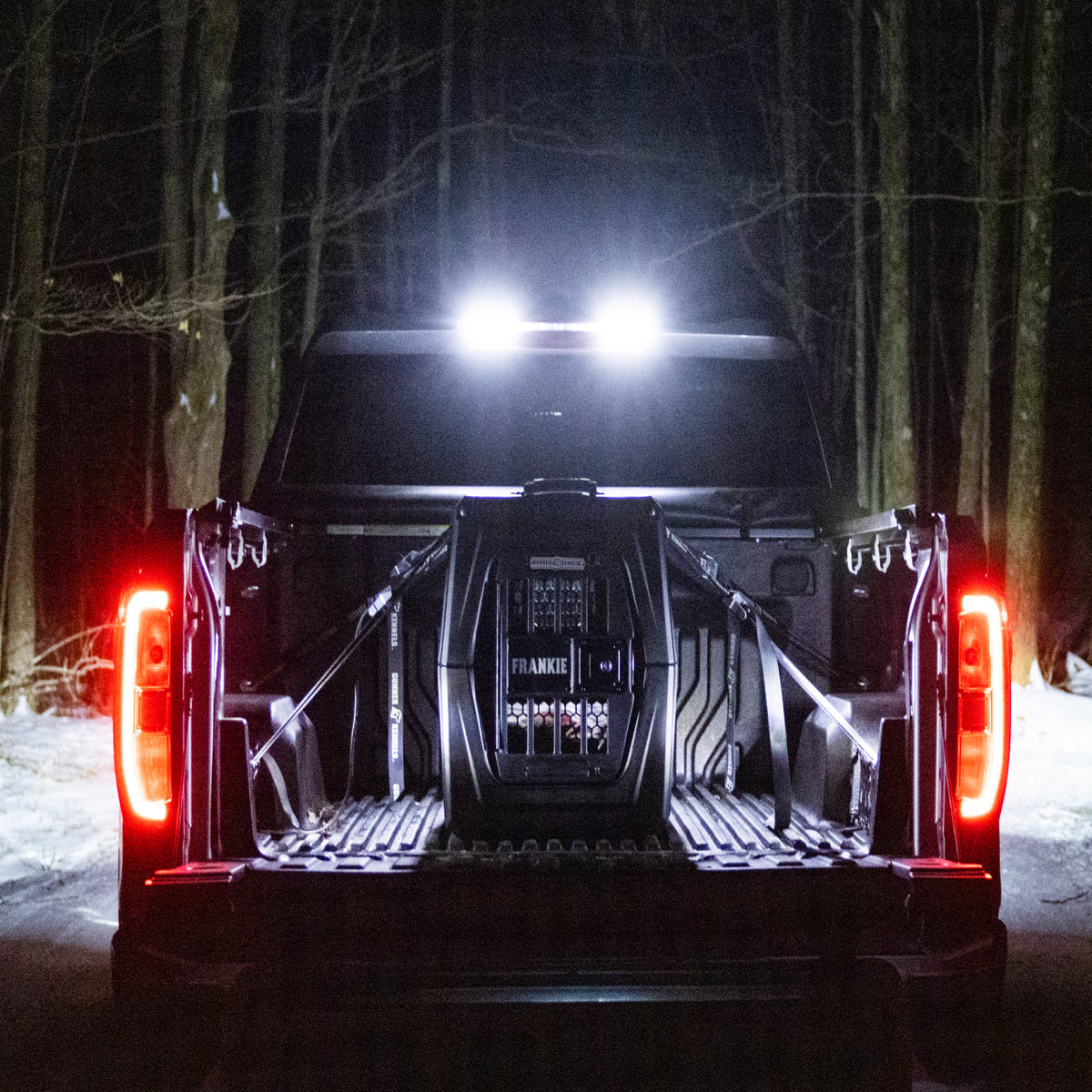 A pickup truck with its tailgate down is parked in a snowy, wooded area at night. Bright overhead lights reveal a GUNNER G1™ KENNEL - BLACK dog crate secured in the truck bed, with red taillights glowing against the dark forest.