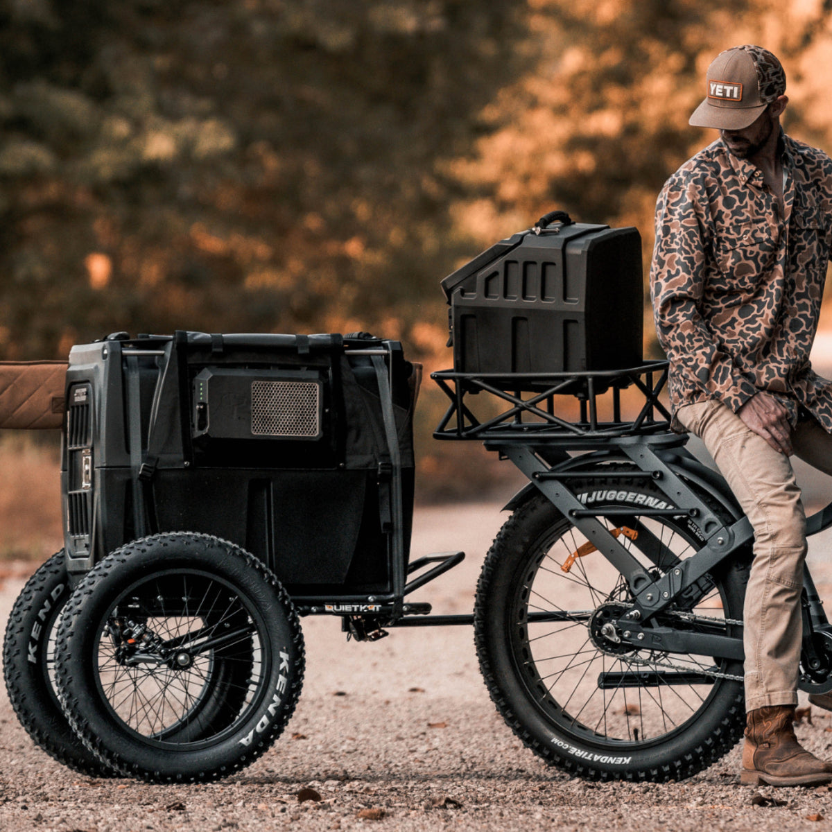 A man in outdoor gear sits on a rugged electric bike with a trailer and a GUNNER G1™ KENNEL - BLACK dog crate attached, parked on a dirt path amid autumn trees.