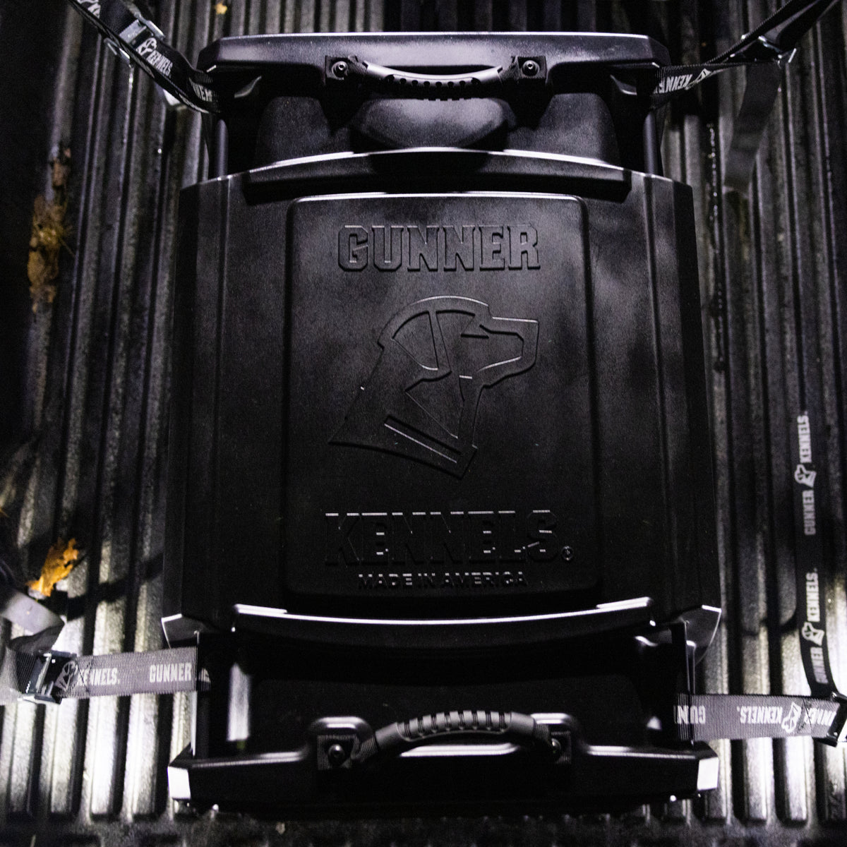 A GUNNER G1™ KENNEL - BLACK sits in a truck bed, secured with Gunner Kennels straps. The crate features an embossed dog head and Made in America.