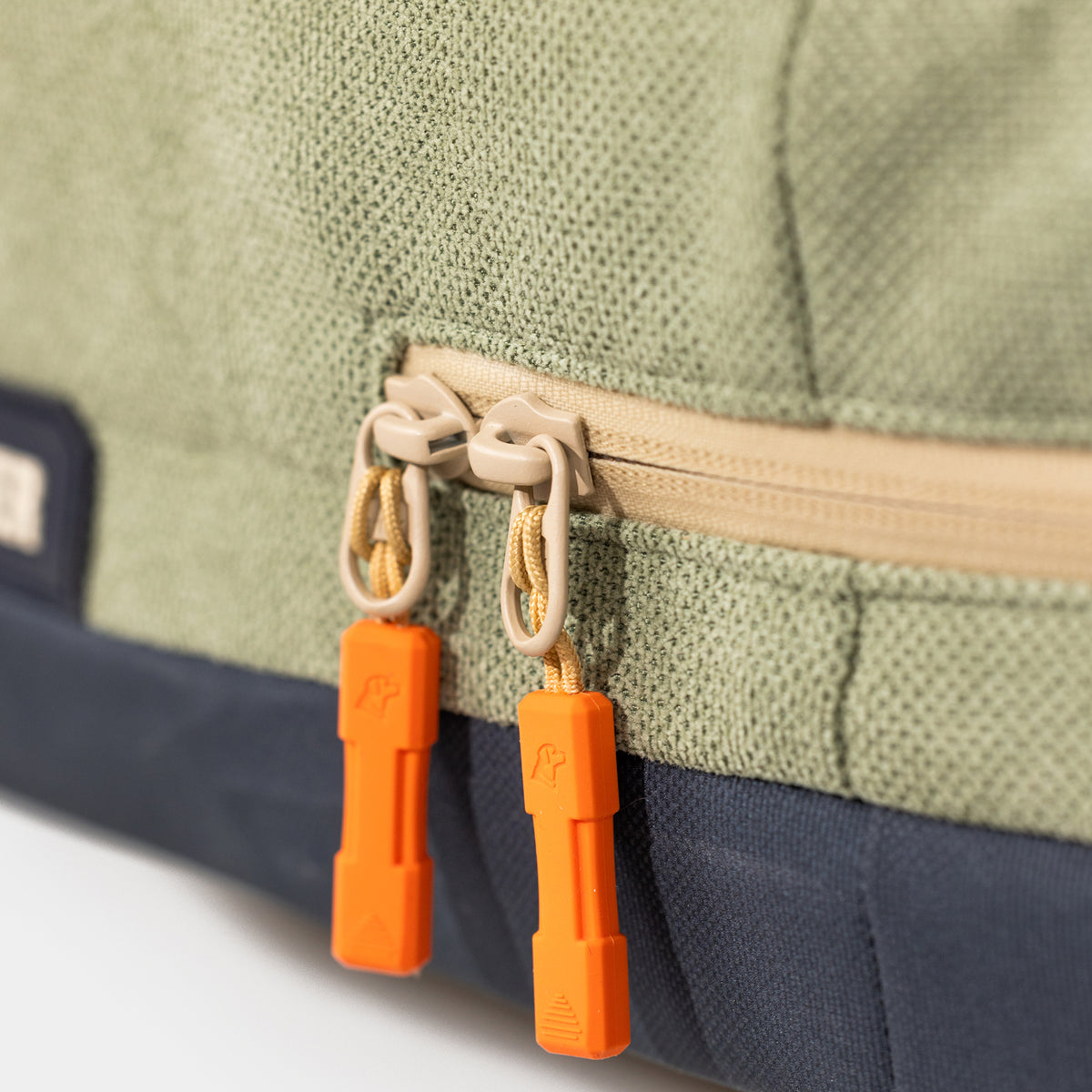 Close-up of the GUNNER HOMESTEAD DOG BED in Treeline Green. The durable fabric shows detailed stitching and a beige zipper with two orange pulls, each embossed with the brand’s logo.
