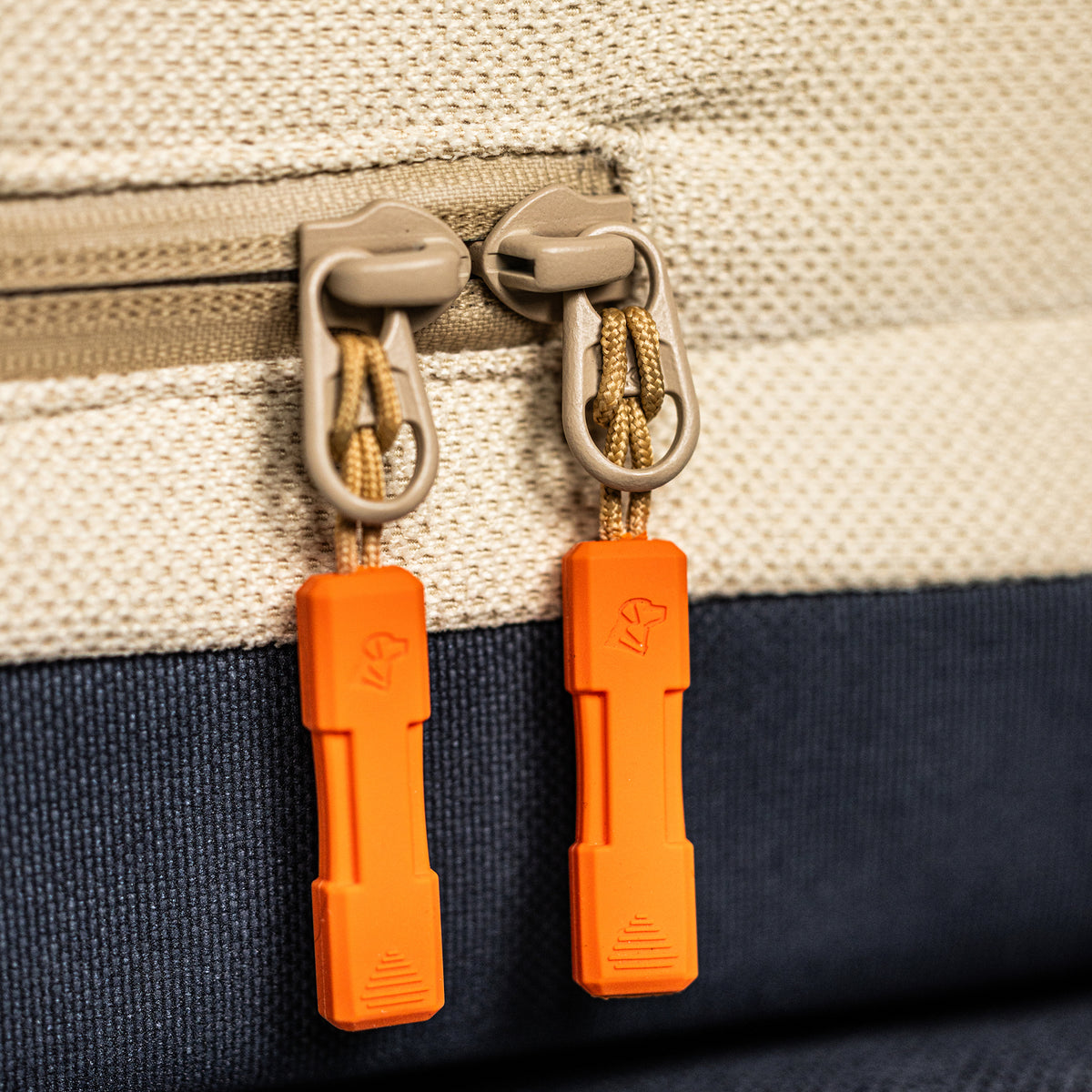 Close-up of two beige zipper pulls with orange grips on the GUNNER HOMESTEAD DOG BED Oak, a durable, textured light-colored fabric bag with a dark blue bottom section.