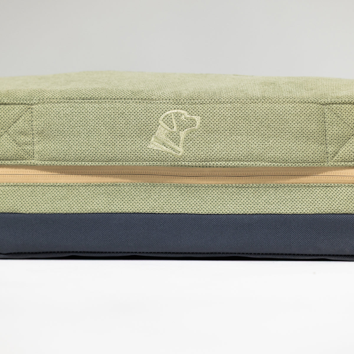 A close-up of the GUNNER HOMESTEAD DOG BED Treeline Green, a rectangular, light green and dark gray cushion with a beige zipper and an embroidered minimalist dog head near the zipper, made from durable, easy-to-clean materials.