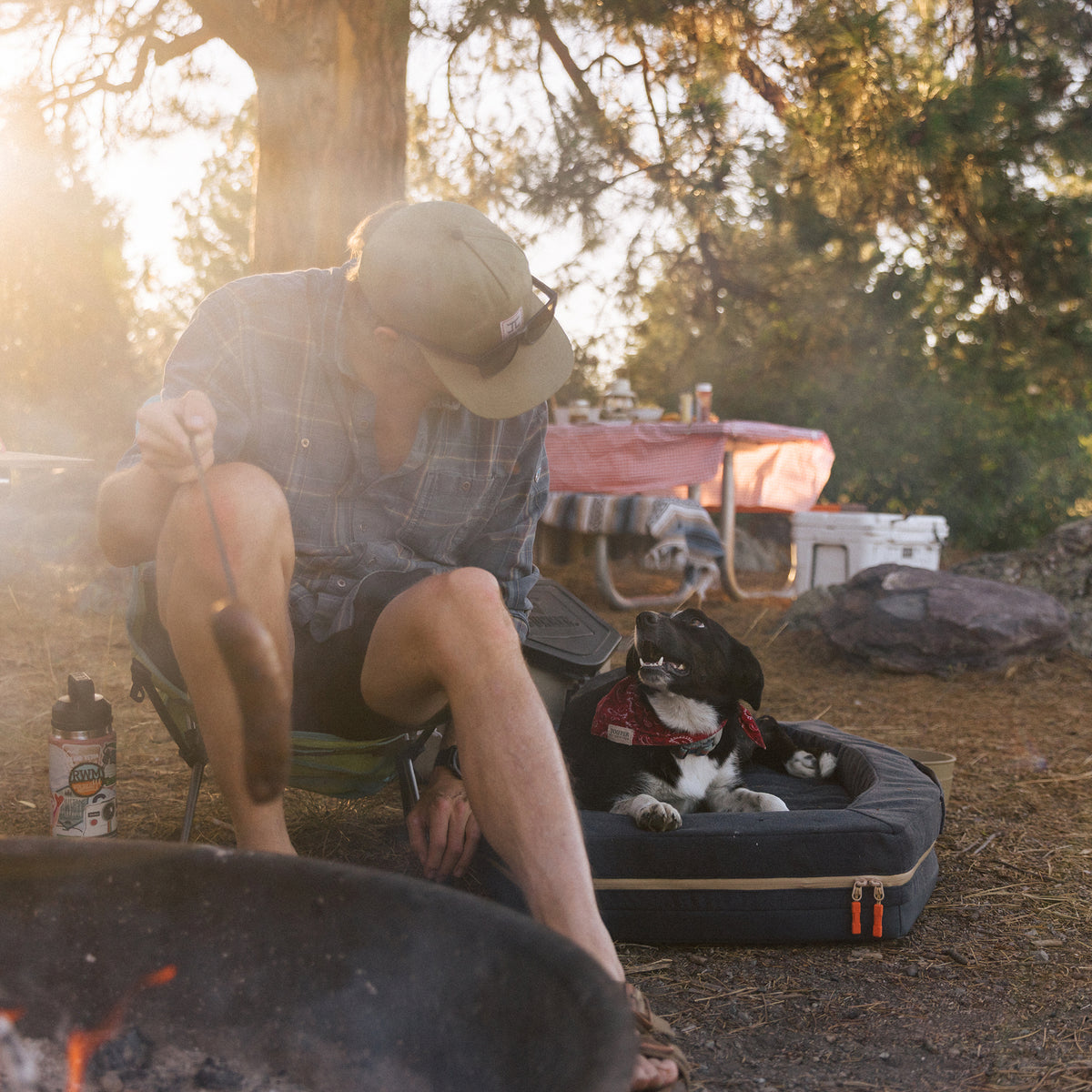 At sunset, a person in a cap and plaid shirt cooks a sausage over a campfire beside their black and white dog—wearing a red bandana—relaxing on the GUNNER HOMESTEAD DOG BED Harbor Blue, surrounded by durable camping gear and trees.