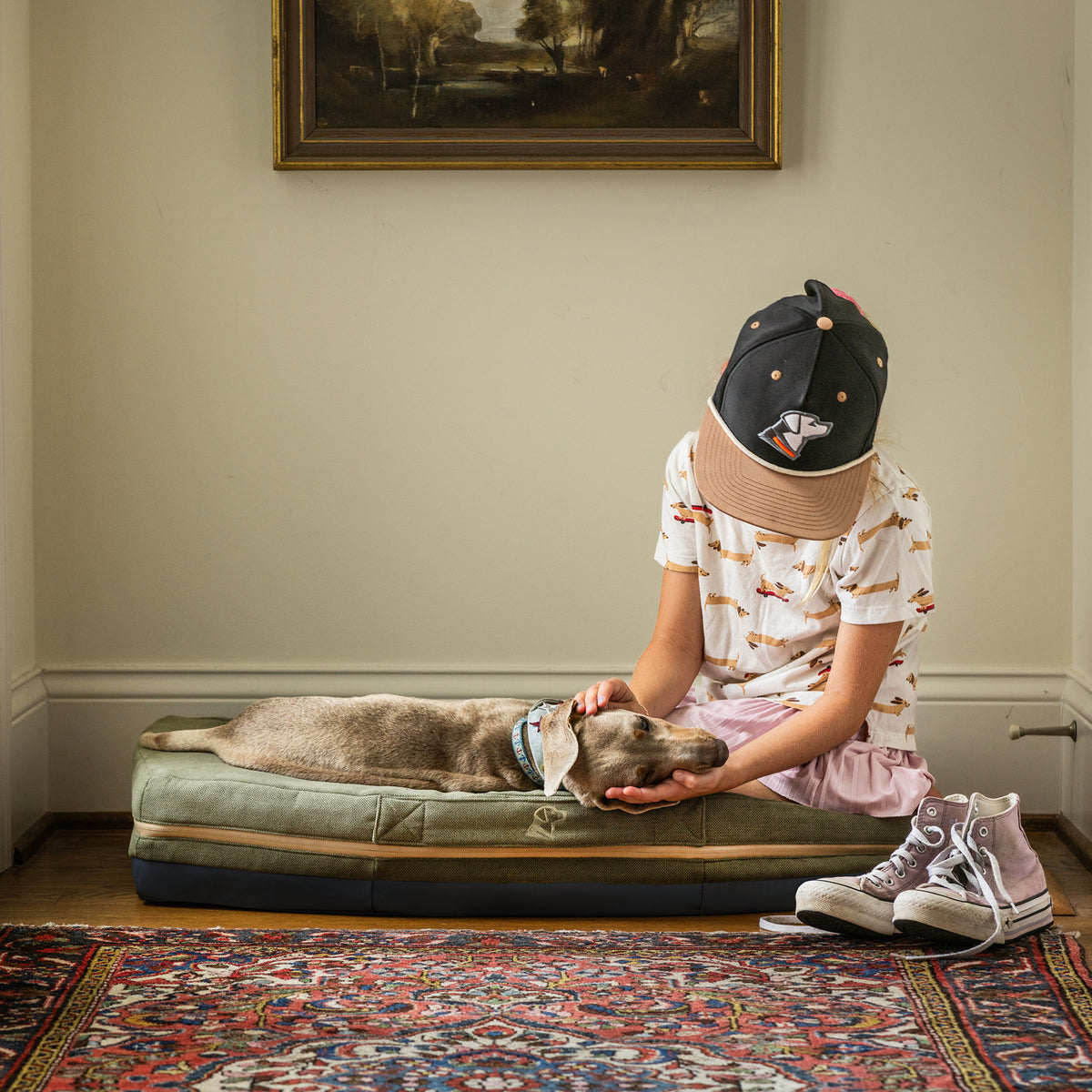 A child in a cap and dog-print shirt sits on the floor, gently petting a gray dog lying on the GUNNER HOMESTEAD DOG BED Treeline Green. A pair of sneakers is nearby, with a framed painting hanging on the wall behind them.