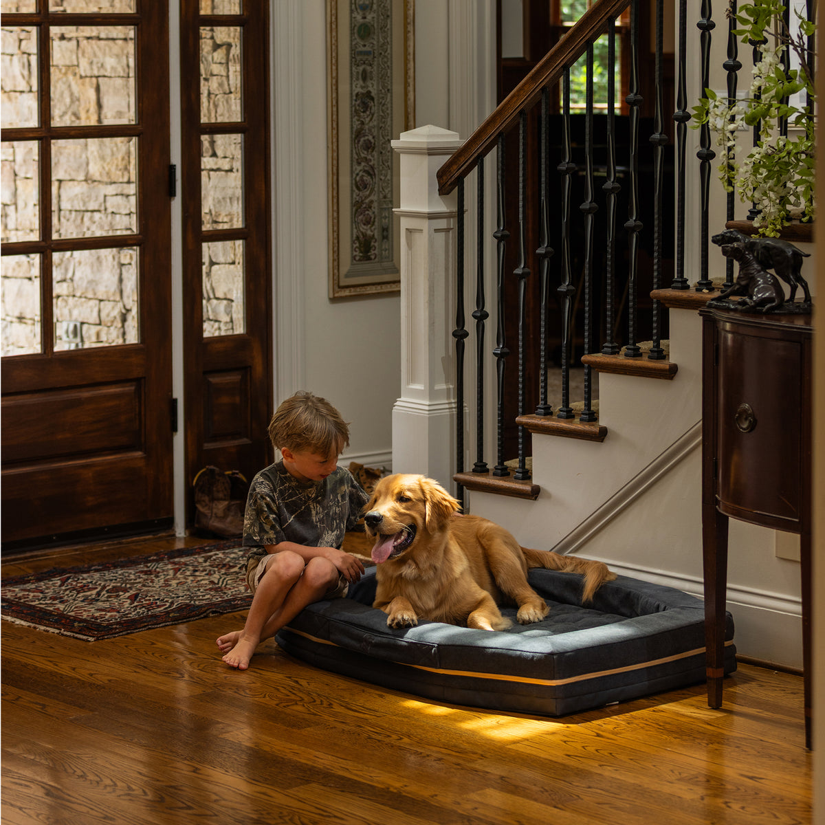 A young boy sits on the floor beside a golden retriever lounging on a GUNNER HOMESTEAD DOG BED in Harbor Blue, set in a warmly lit, elegant entryway with wooden floors and a staircase in the background.