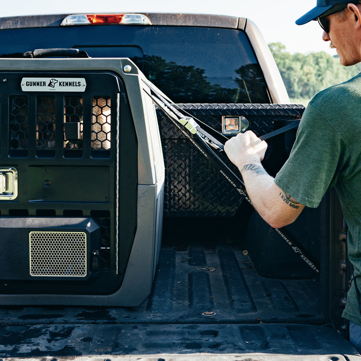 A person in a green shirt straps the GUNNER G1™ KENNEL - GUNMETAL in a pickup truck bed with a tie-down. Trees in the background emphasize how this reliable crate can help protect dogs on the road.