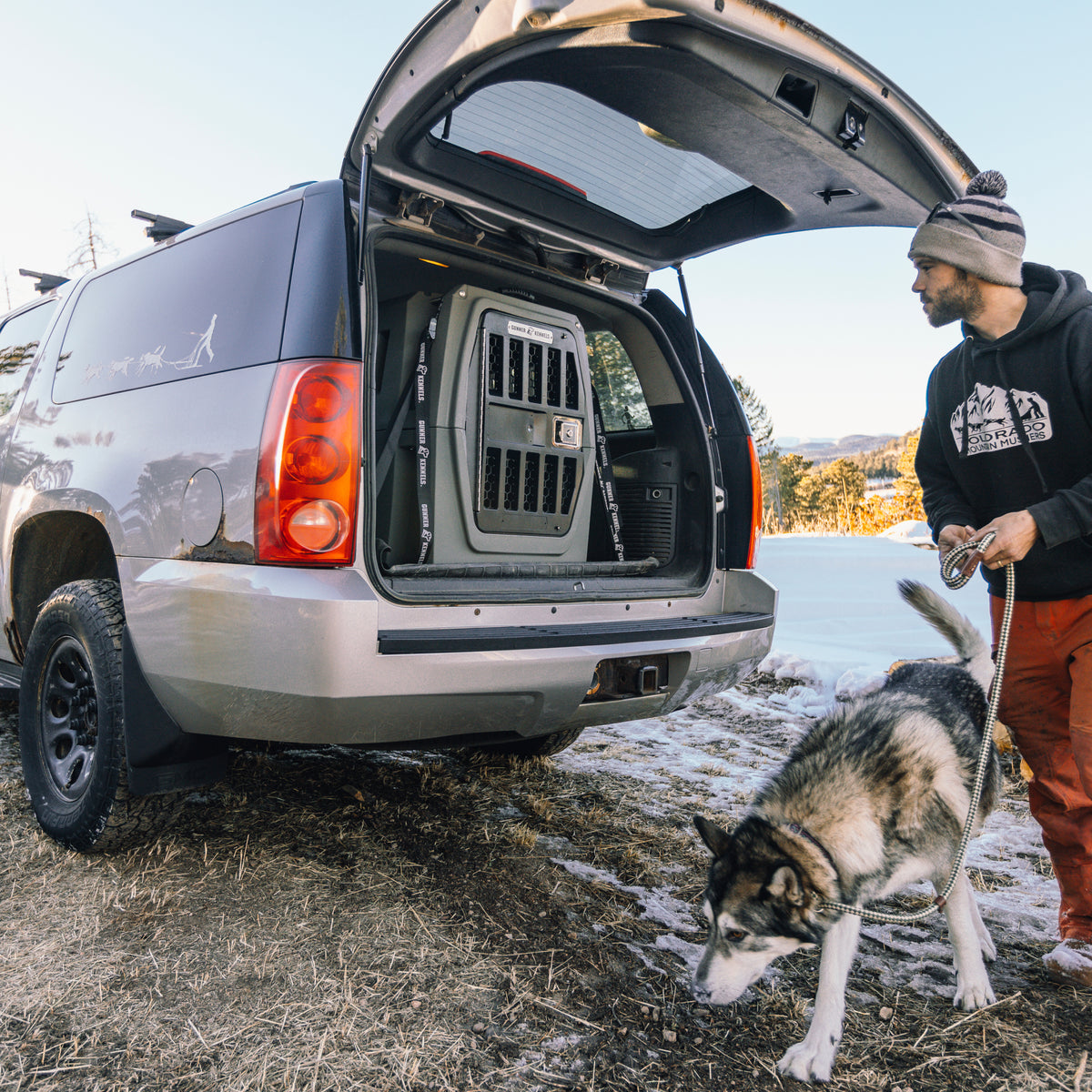 A man in a beanie and hoodie stands with his leashed husky next to an SUV with its back open, showing a GUNNER G1™ KENNEL - GUNMETAL inside. The chilly outdoor setting includes some snow on the ground.