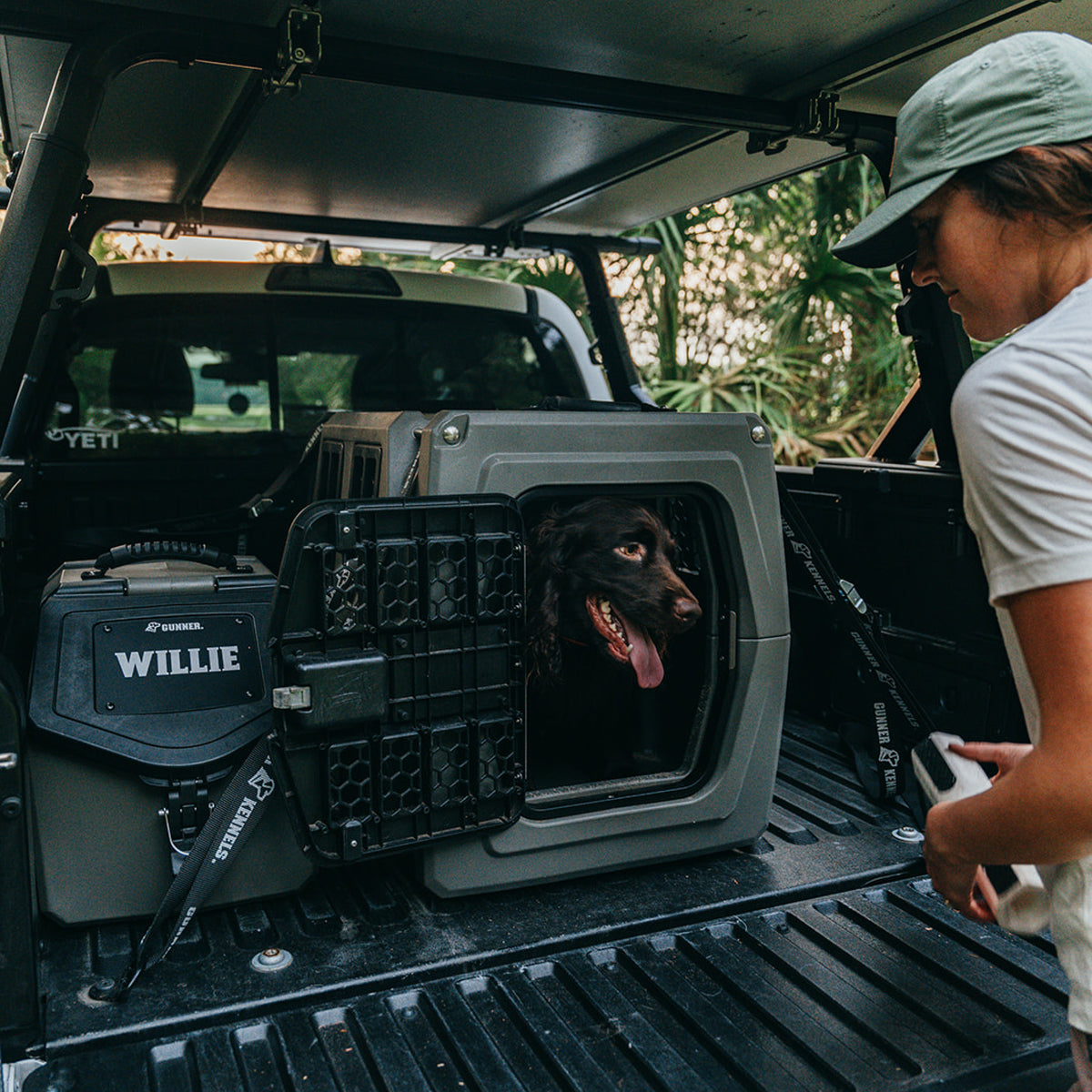 A woman stands by an open truck bed where her dog, Willie, sits inside the GUNNER G1™ KENNEL - GUNMETAL, looking out with its tongue hanging as greenery fills the outdoor scene.