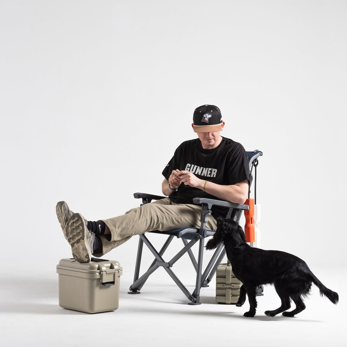 A man in a black GUNNER T-shirt and a HUNT SOLIDS Rope Hat sits cross-legged on a folding chair next to beige storage boxes, with a black dog standing in front. The background is plain white.