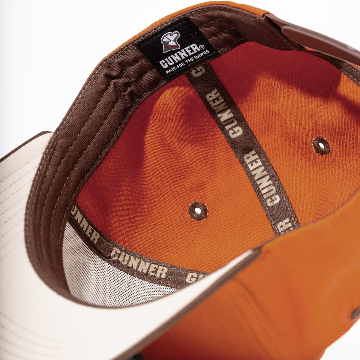 Close-up of the HUNT SOLIDS Snapback by GUNNER: orange/white cap with brown trim, labeled seams reading “GUNNER,” and a black tag stating “GUNNER MADE FOR THE GUN DOGS. Features Perfect Fit snapback design.