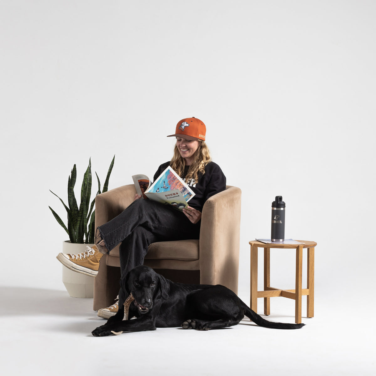 A woman in a tan armchair reads a colorful magazine, wearing a GUNNER HUNT SOLIDS Snapback and sneakers. Her black dog lies nearby. A plant and water bottle rest on a small table against a plain white background.