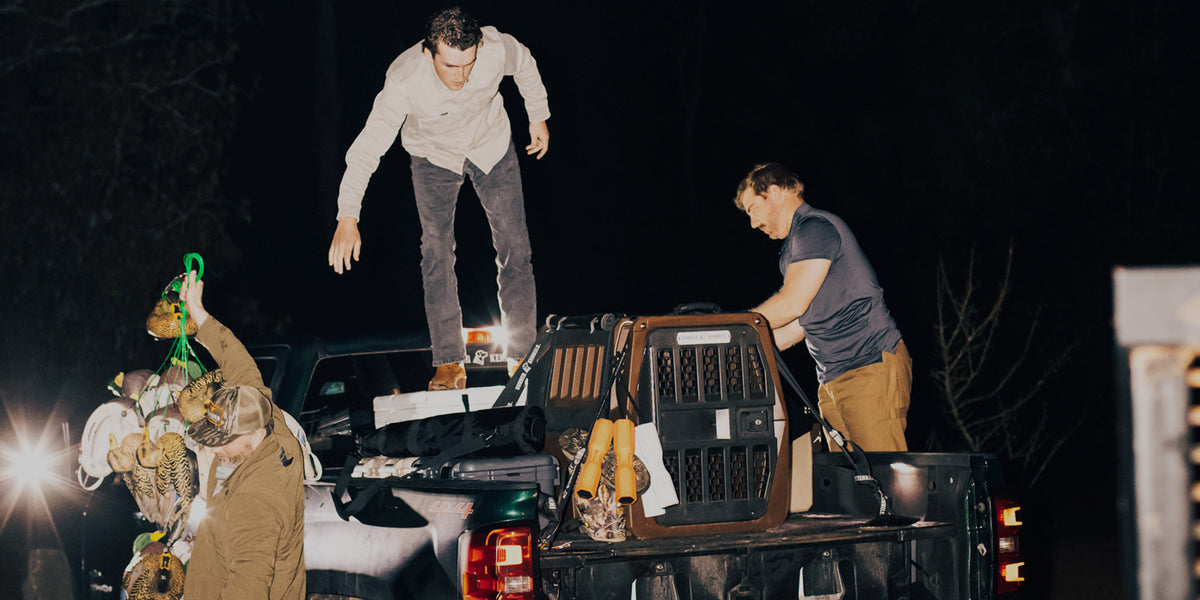 Three people load supplies and a dog kennel onto the back of a pickup truck at night. One stands on the truck bed, another hands up items, while a third organizes equipment. The scene is lit by vehicle headlights.