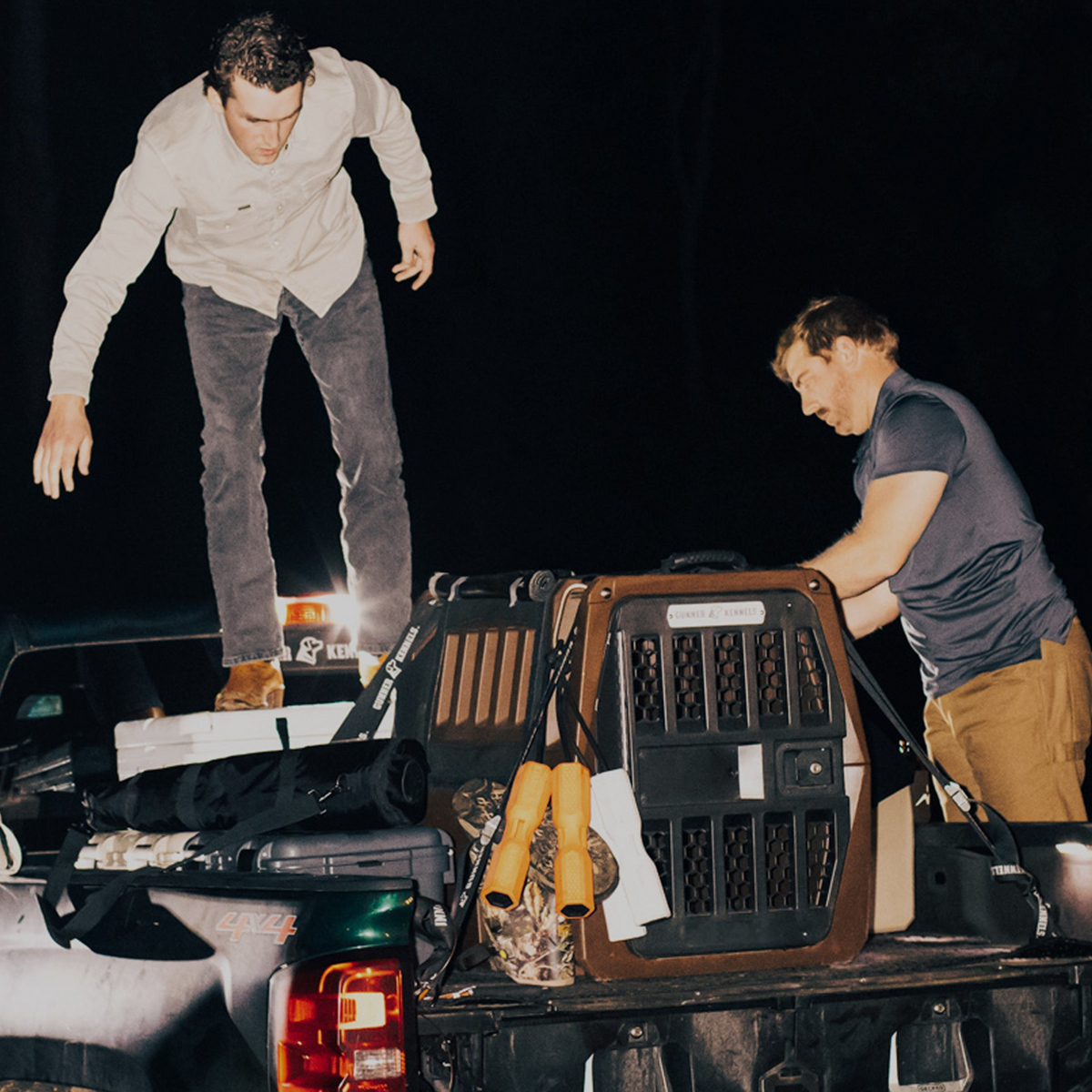 Two men unload items, including crates and gear in Flyway colors, from a pickup at night. One stands on the truck bed while the other arranges objects beside a large GUNNER G1™ KENNEL - SUSQUEHANNA.