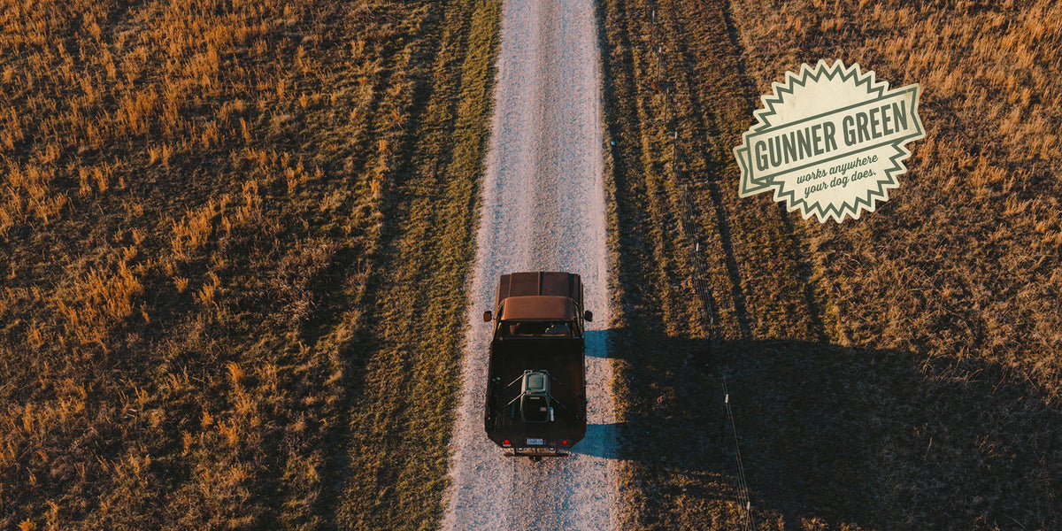 Aerial view of a truck driving down a gravel road surrounded by grassy fields. A vintage-style badge in the upper right corner reads “GUNNER GREEN,” perfect for those seeking dependable outdoor gear for every working dog adventure.