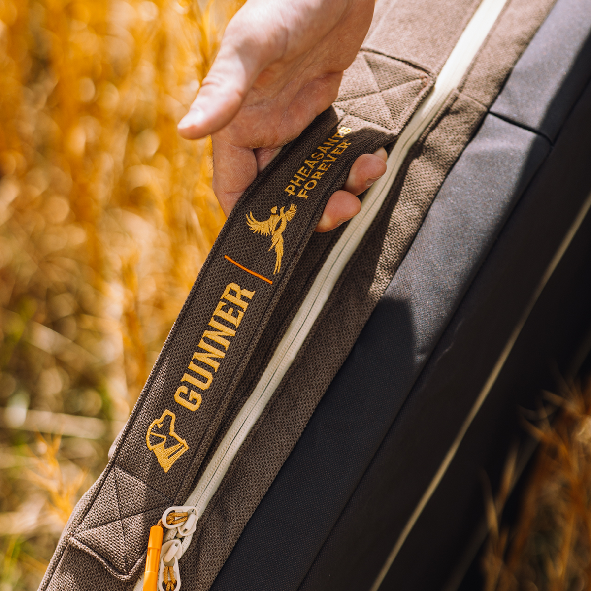 A hand grips a durable HOMESTEAD DOG BED in Special-Edition Pheasant Brown by GUNNER, with Pheasants Forever collaboration embroidered in gold. Golden reeds blur softly around the bed.