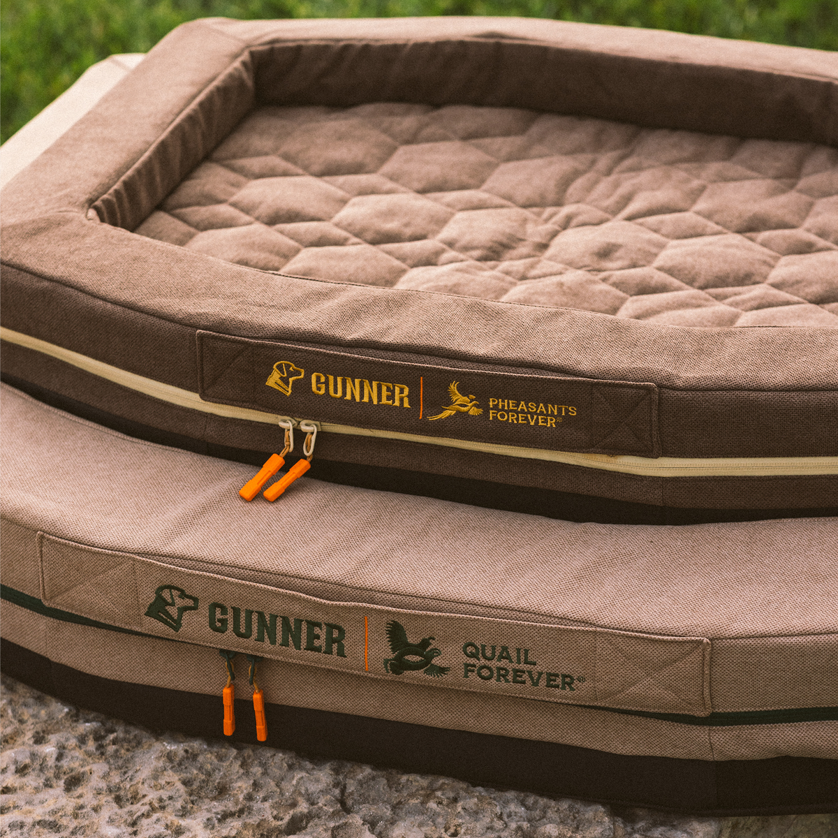 Two brown, rectangular GUNNER HOMESTEAD DOG BEDs in Quail Tan are stacked outdoors; the top features GUNNER and Pheasants Forever logos, while the bottom shows GUNNER and Quail Forever Tan logos.