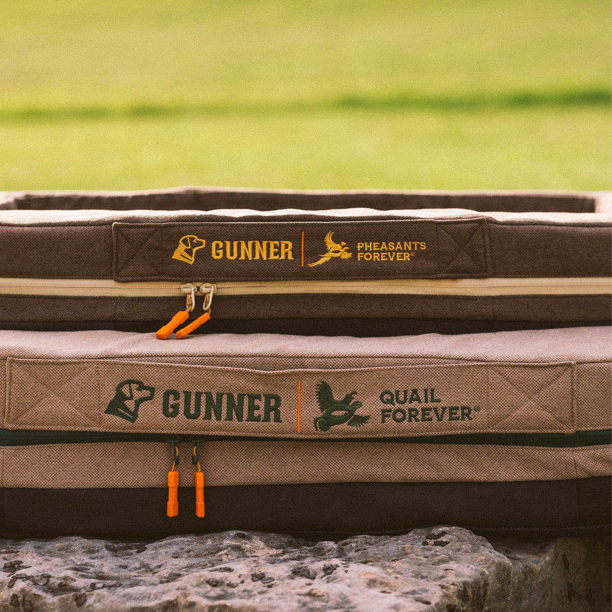Two brown and tan rectangular dog beds on a stone surface, designed to complement your dog kennel or crate, featuring GUNNER branding and logos for Pheasants Forever and Quail Forever with orange zipper pulls, outdoors with grass in the background.