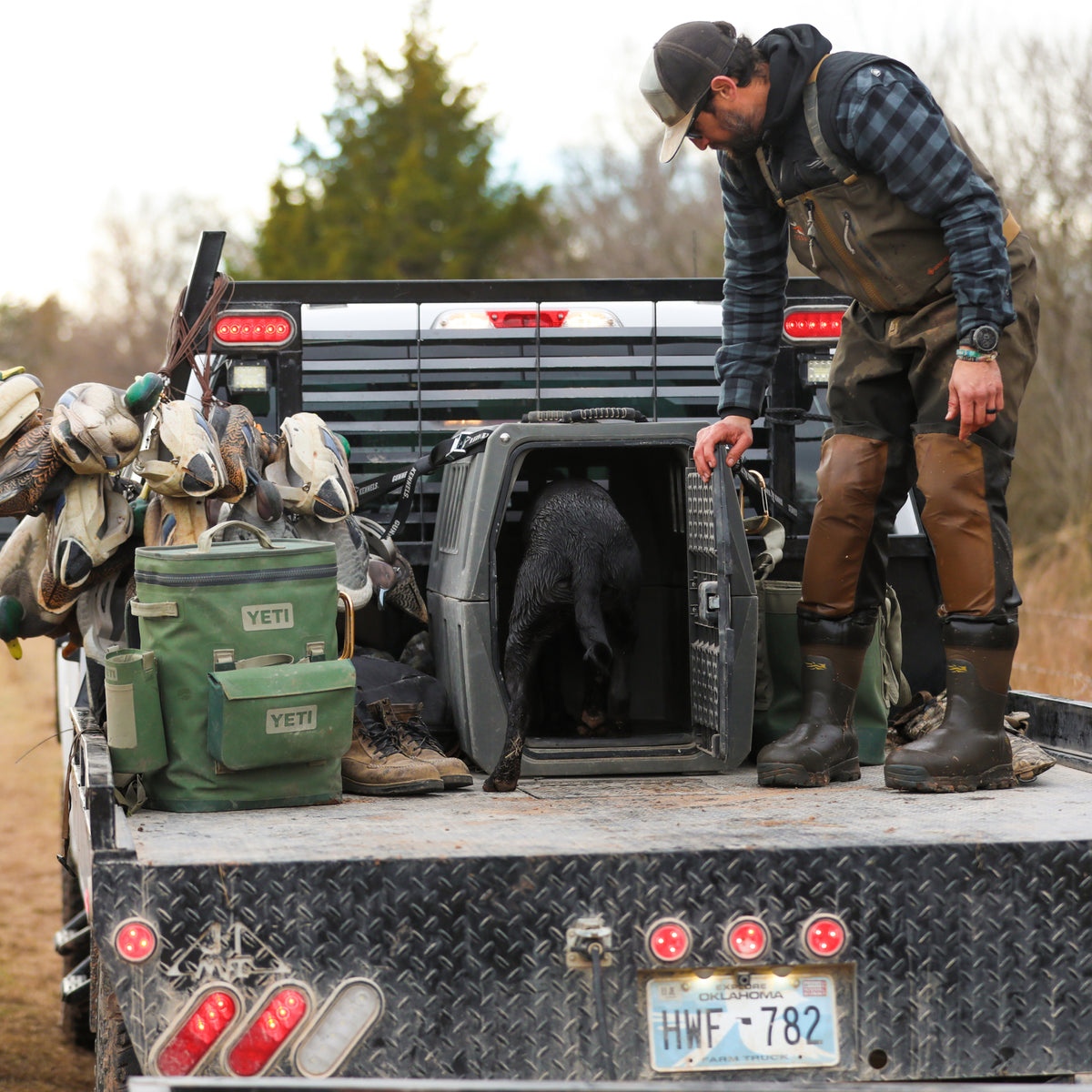 A man in waders stands on a truck bed loaded with hunting gear, opening a GUNNER G1™ KENNEL - MOSSY OAK BOTTOMLAND GUNMETAL for his black dog. Duck decoys, bags, and boots sit nearby under an overcast sky and trees.