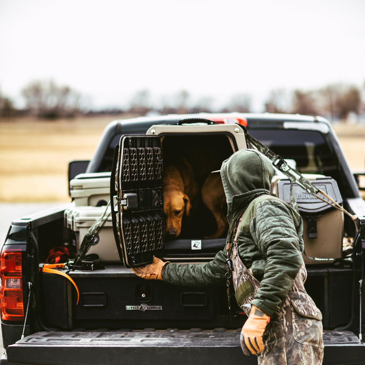 Wearing outdoor gear, a person opens a GUNNER G1™ KENNEL - TAN/SHADOW GRASS in the back of a pickup. Two brown dogs are inside the camo kennel, with rural fields visible in the background.
