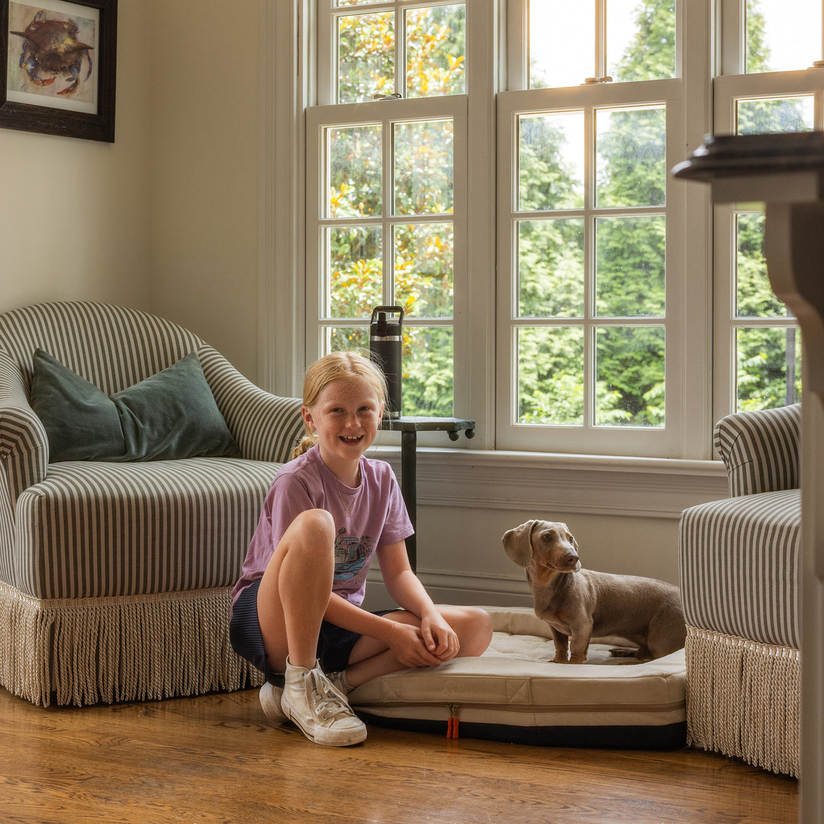 A child with blonde hair and a purple shirt sits indoors next to a small brown dog on the GUNNER HOMESTEAD DOG BED Oak, with two armchairs and large windows behind them.