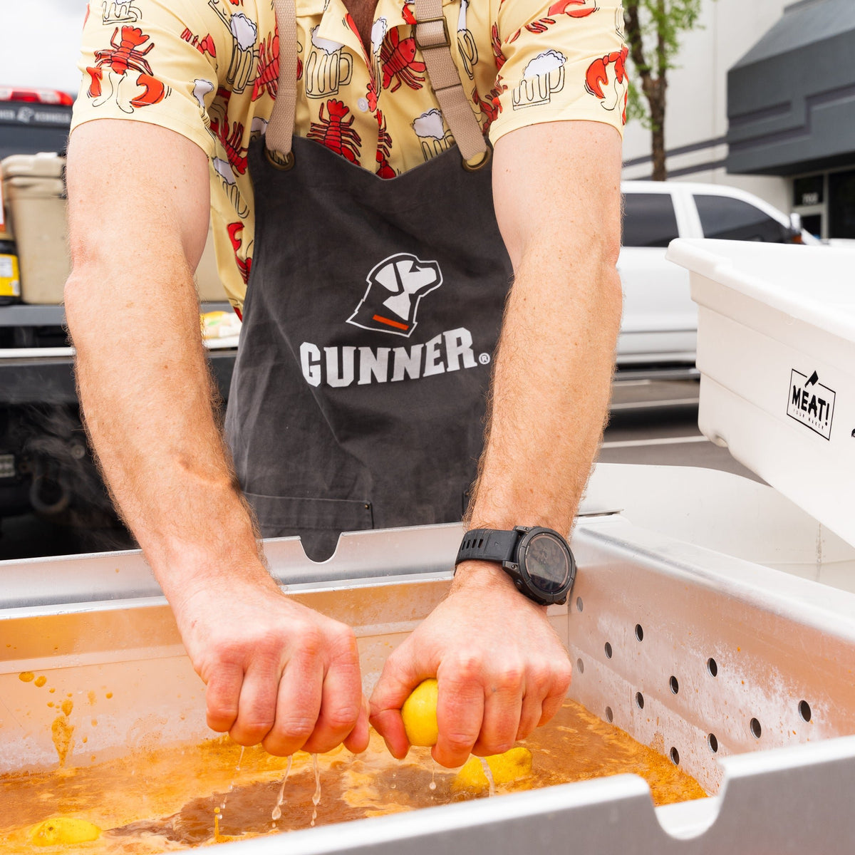 Wearing a patterned shirt and the GUNNER Apron by GUNNER, a person squeezes lemons into a large metal container of orange liquid outdoors, possibly prepping food for tailgates or crawfish boils.