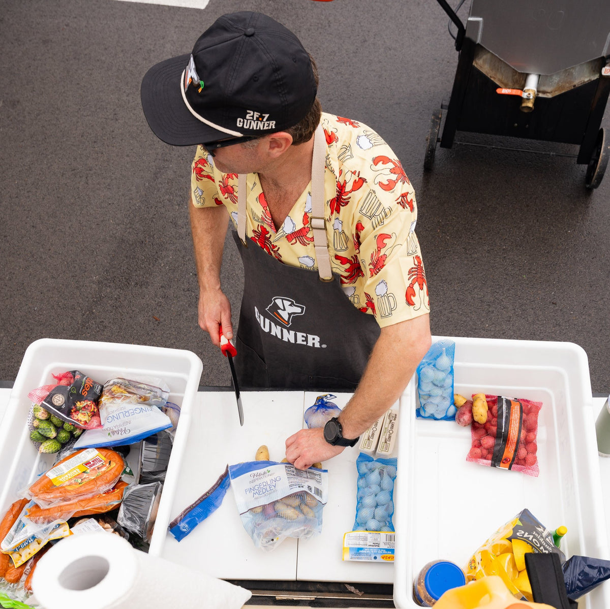 A man in a patterned shirt and cap wears a GUNNER Apron by GUNNER while chopping potatoes on a white table outdoors, surrounded by colorful potato bags—prepping ingredients for crawfish boils or tailgates at a bustling market.