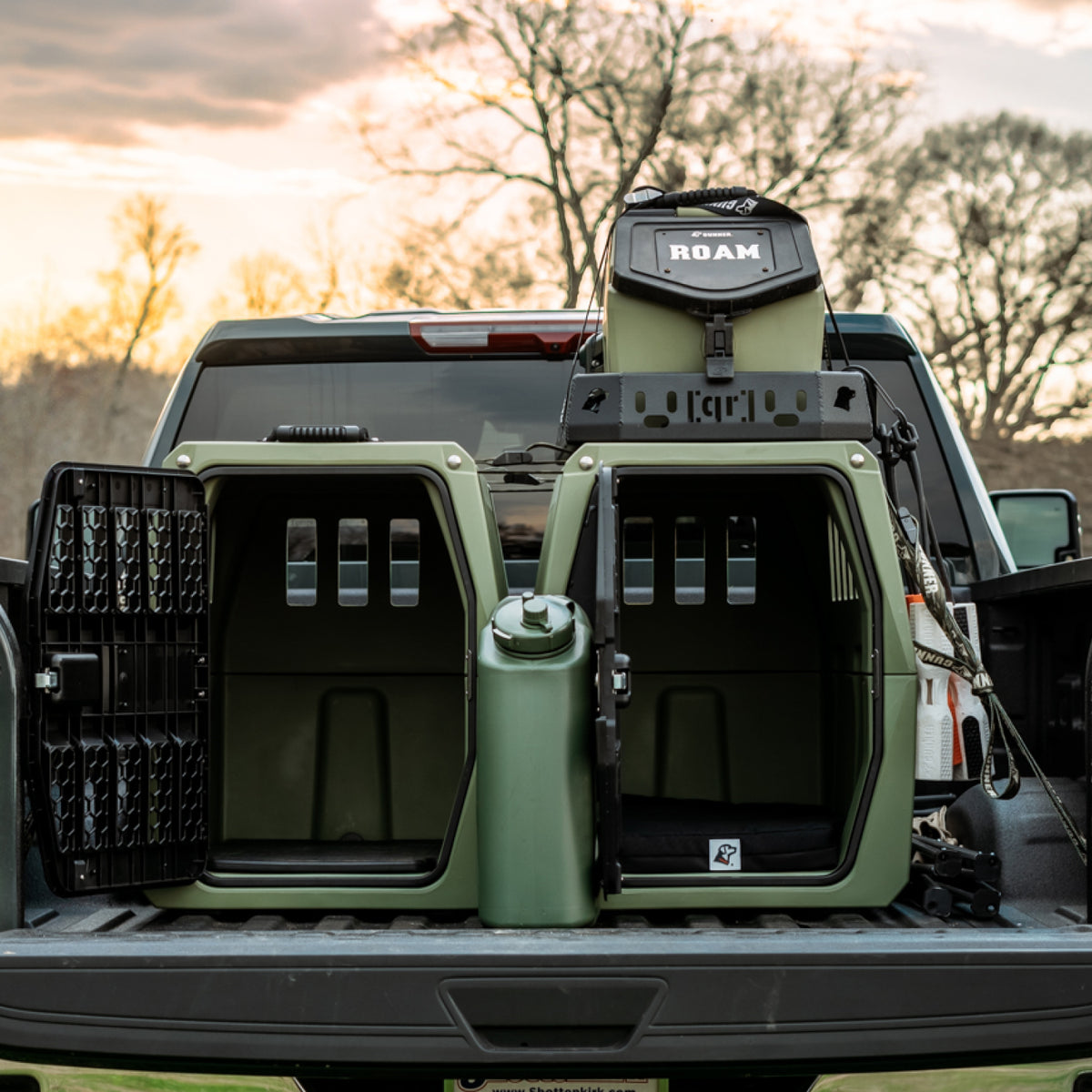 Two green dog kennels with open doors are placed in the back of a pickup truck. A green water container and a black storage bag are nearby. Trees and a cloudy sky are in the background.