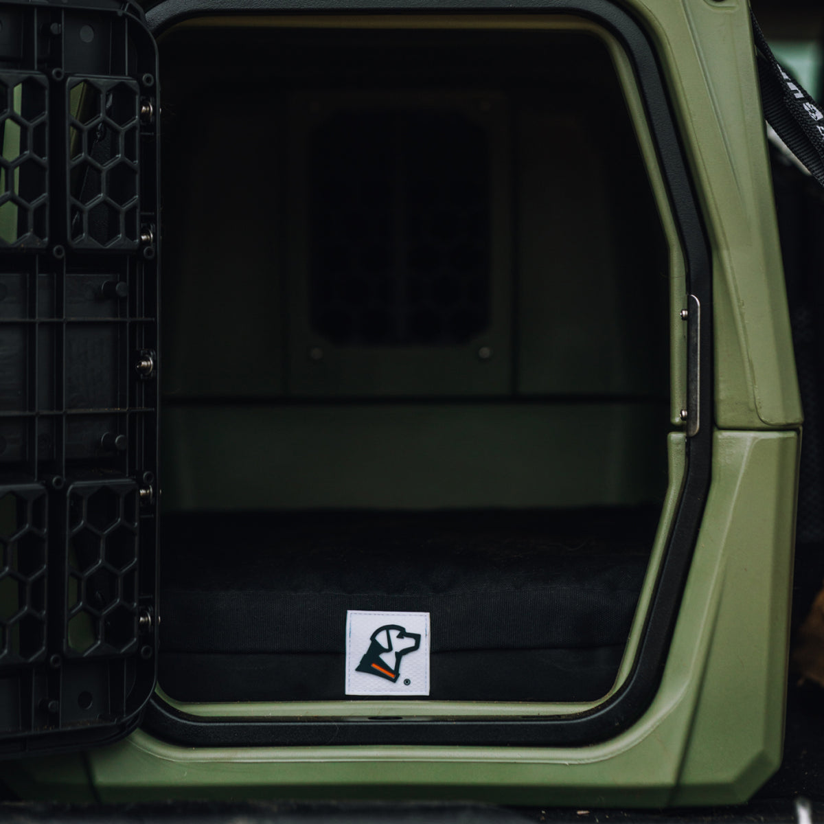 An open green dog crate with a black cushion inside, featuring a small logo of a dog’s head on the cushion—ideal outdoor gear for your working dog.
