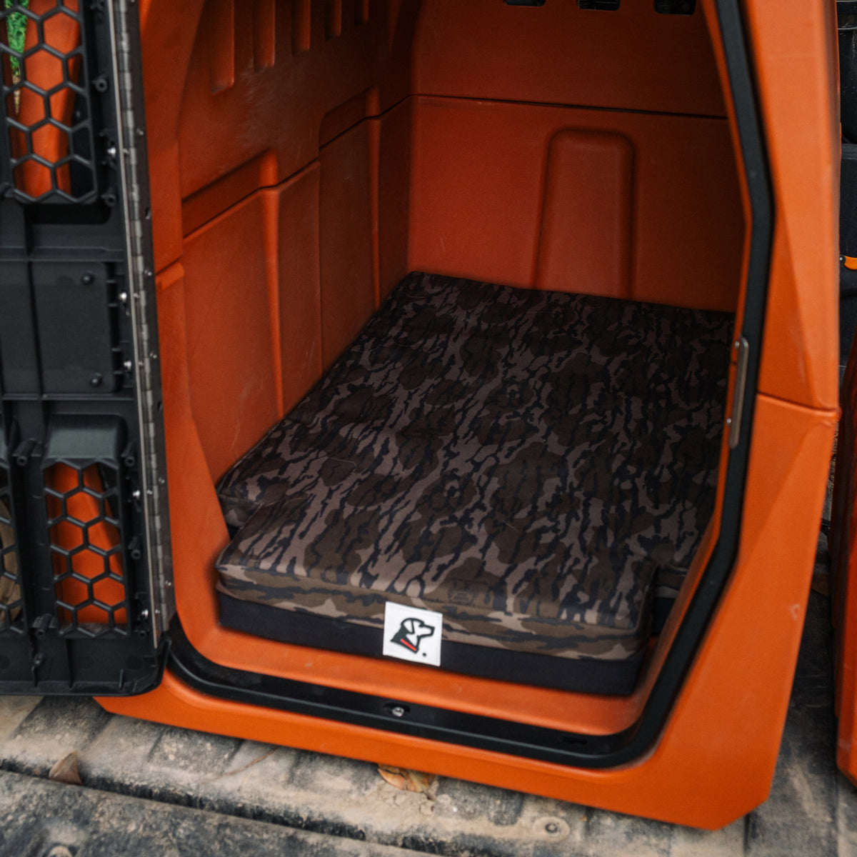 Open orange kennel with a dark camouflage-patterned dog bed inside, viewed from the side. Designed as reliable outdoor gear for any working dog, the bed has a small logo on the corner. The kennel is empty and sitting on a textured surface.