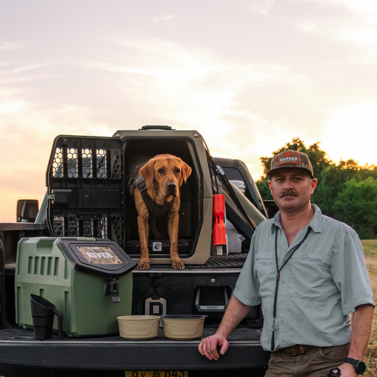 A man in outdoor clothing stands beside a truck with a brown dog in a kennel on the truck bed. The scene is outdoors at sunset, with pet supplies on the truck.