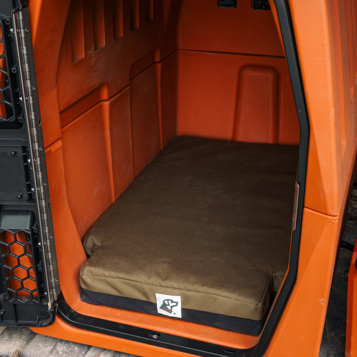 An orange dog kennel with a thick, olive green cushion inside. The kennel door is open, revealing the interior and the cushion with a small white logo in the front corner—perfect for showcasing your pup’s favorite outdoor gear.