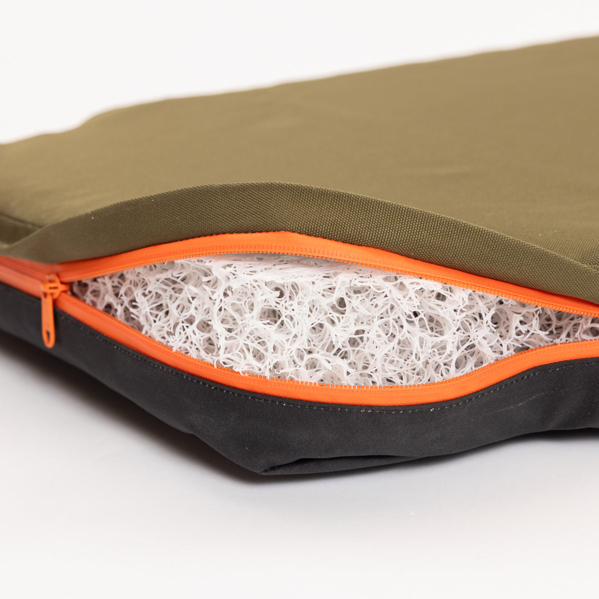 A close-up of a green and black cushion—reminiscent of rugged outdoor gear—with an orange zipper partially open, revealing a white, tangled mesh filling inside.