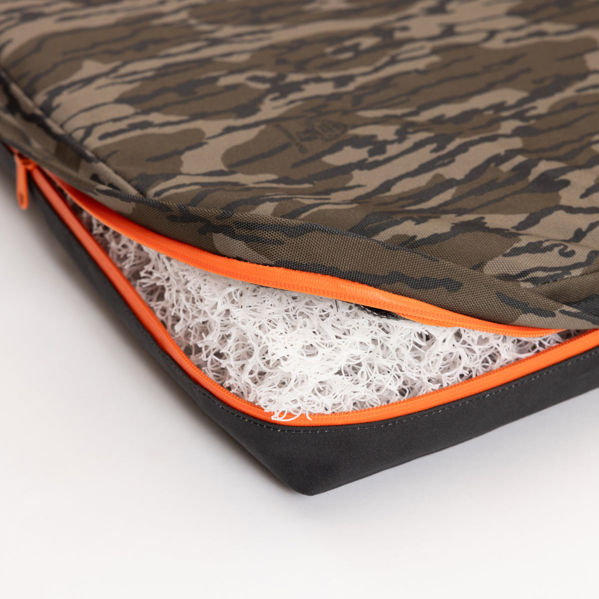 A close-up of a camouflage-patterned cushion, perfect for outdoor gear or a hunting dog’s crate, with an orange zipper partially open to reveal a white, curly, mesh-like filling inside.