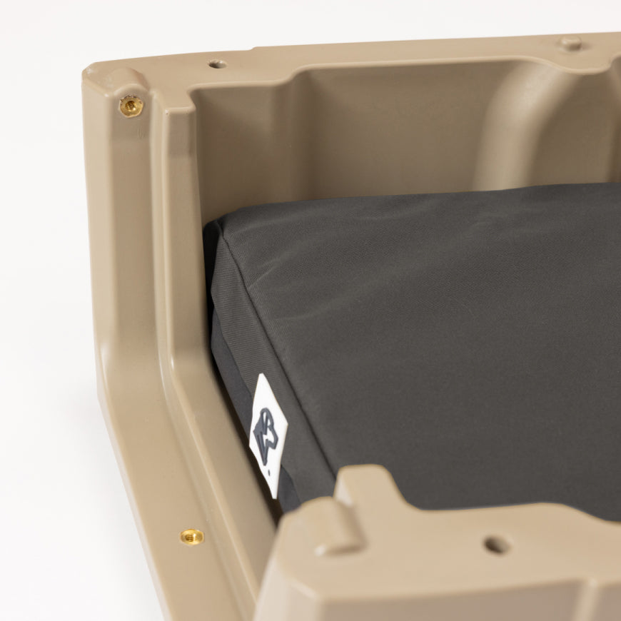 Close-up of a beige plastic dog crate frame holding a thick, dark gray cushion with a small white tag attached, set against a plain white background.