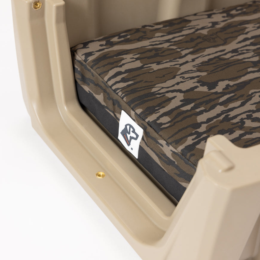 Close-up of a tan plastic dog crate with a brown and black camouflage cushion inside. A small logo tag is attached to the cushion near one corner.