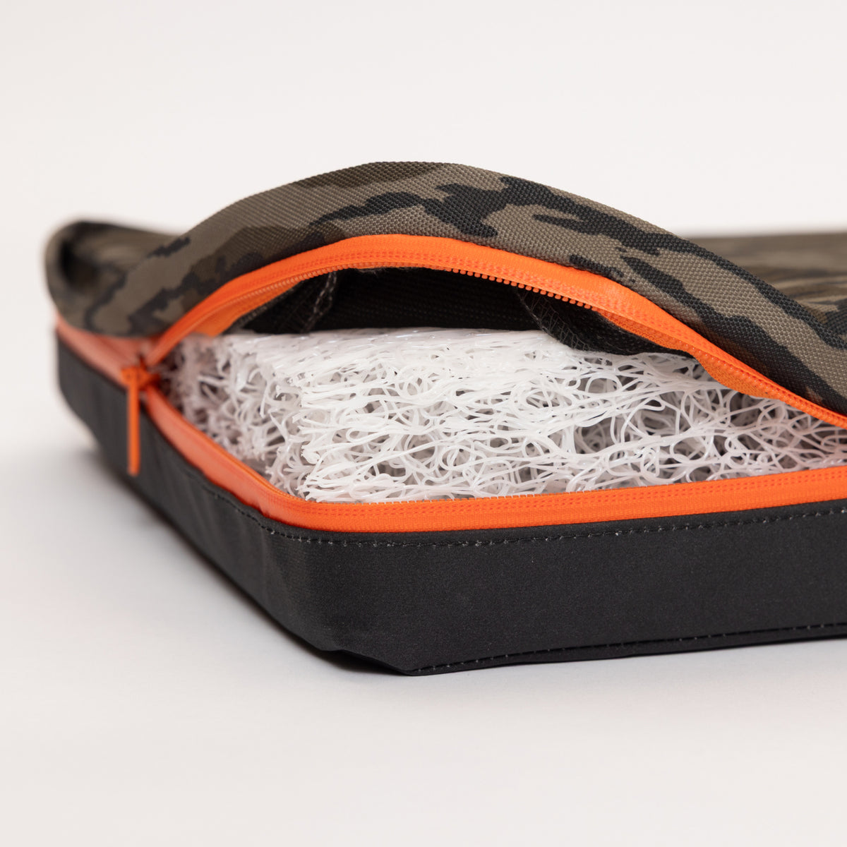 A close-up of a zippered case with a camouflage pattern and orange zipper, partially open to reveal a white, mesh-like cushion or pad inside—perfect for adding comfort to your working dog’s kennel or dog crate.