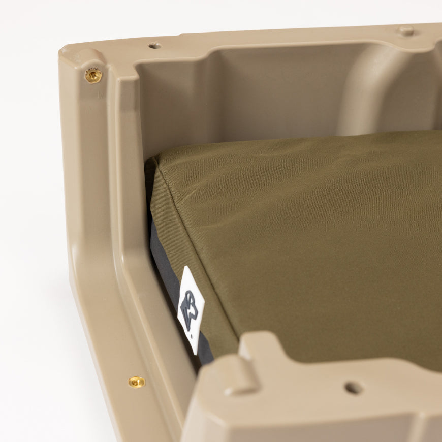 A close-up of a tan plastic dog crate with a green cushion inside. The cushion has a small white tag with a black logo on the corner.