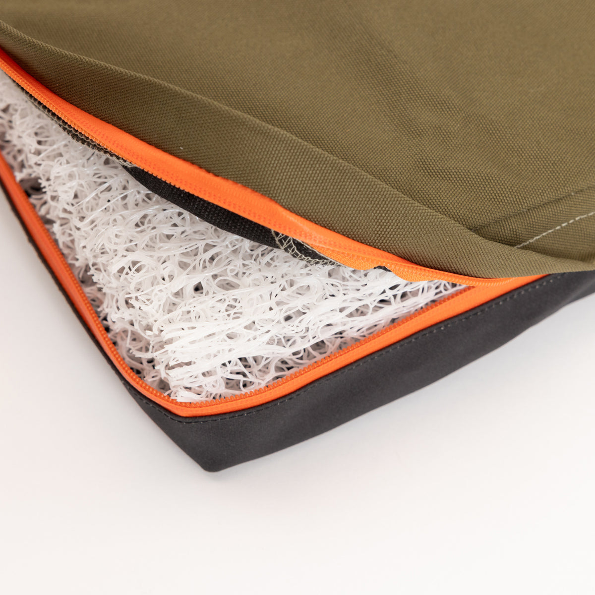 A close-up of an opened cushion or mattress with an olive green and black cover, designed for use in a dog crate or as outdoor gear, showing a white, mesh-like inner material and an orange zipper.