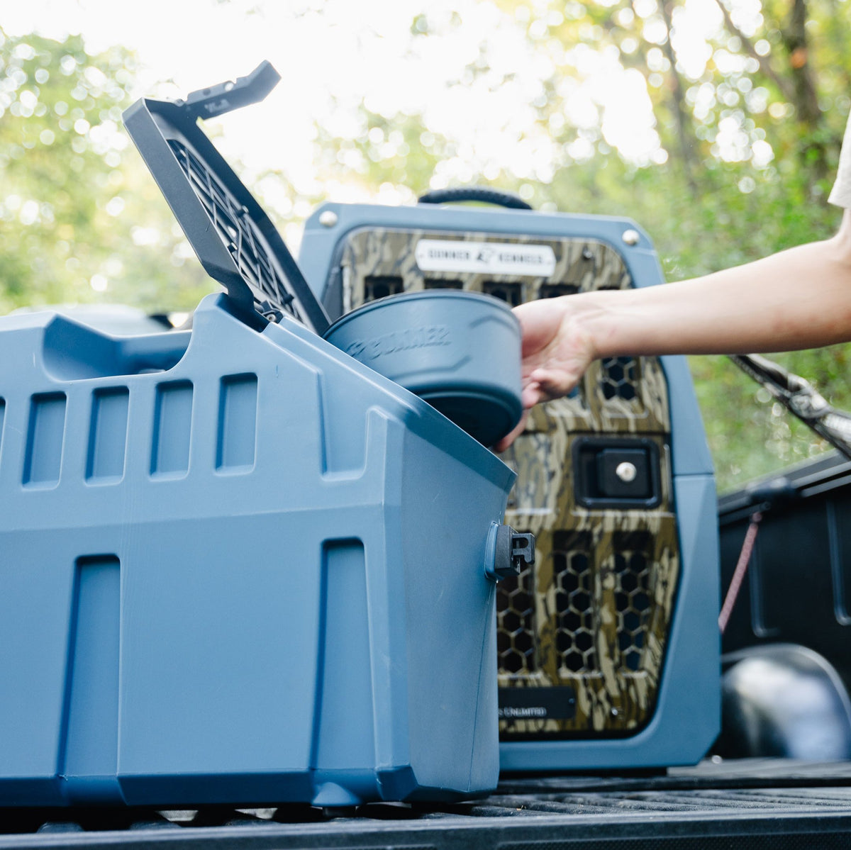 A person places a water bowl into a GUNNER FOOD CRATE - Ducks Unlimited Blue, while an airtight dog food container sits in another camouflaged crate outdoors among trees.