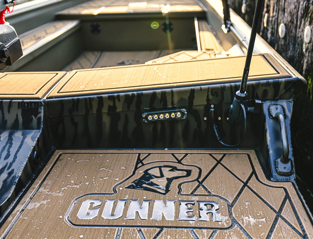 Close-up of a boat’s deck with GUNNER and a dog’s head logo on textured brown flooring, rod holders, and LED lights near a wooden dock—a dog kennel is also secured on the deck for your companion’s comfort.