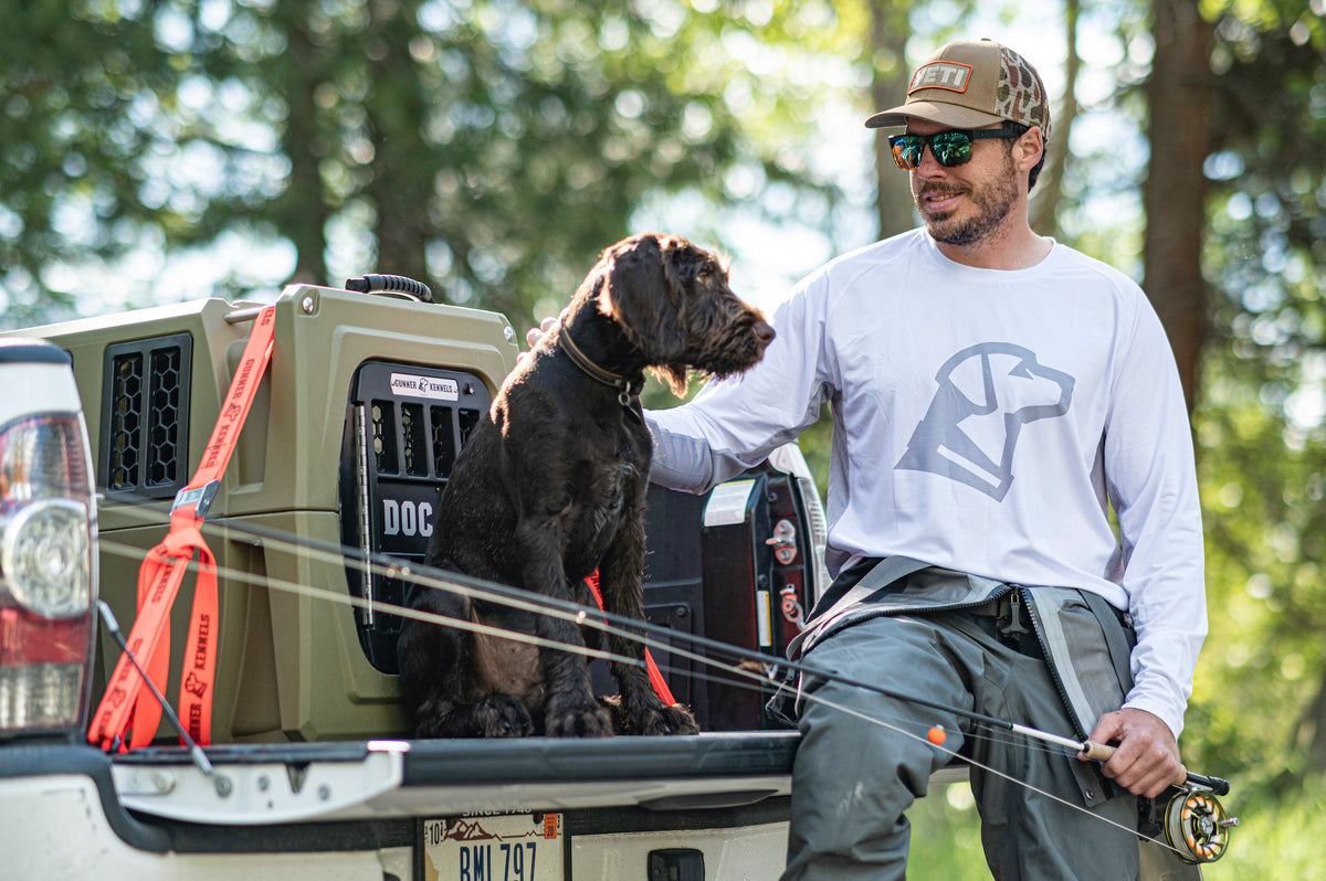 A man wearing sunglasses and a hat sits on the tailgate of a truck with fishing rods, petting his hunting dog sitting beside him near a dog crate, outdoors with trees in the background.