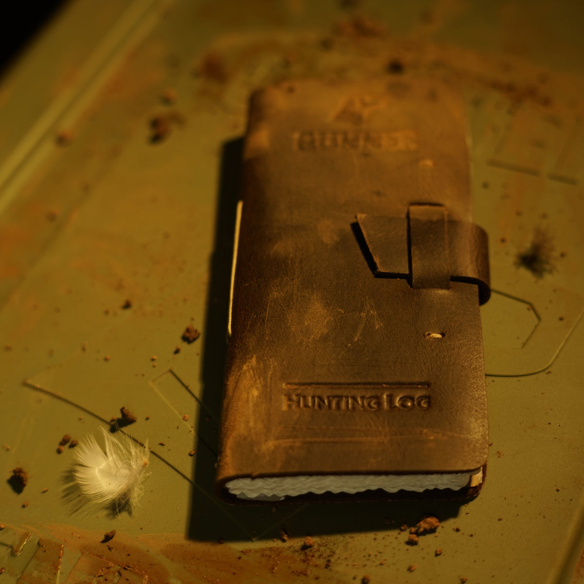 The GUNNER Hunting Log, a worn leather journal with a strap, rests on a dusty green surface beside a white feather—evoking the spirit of outdoor hunts.